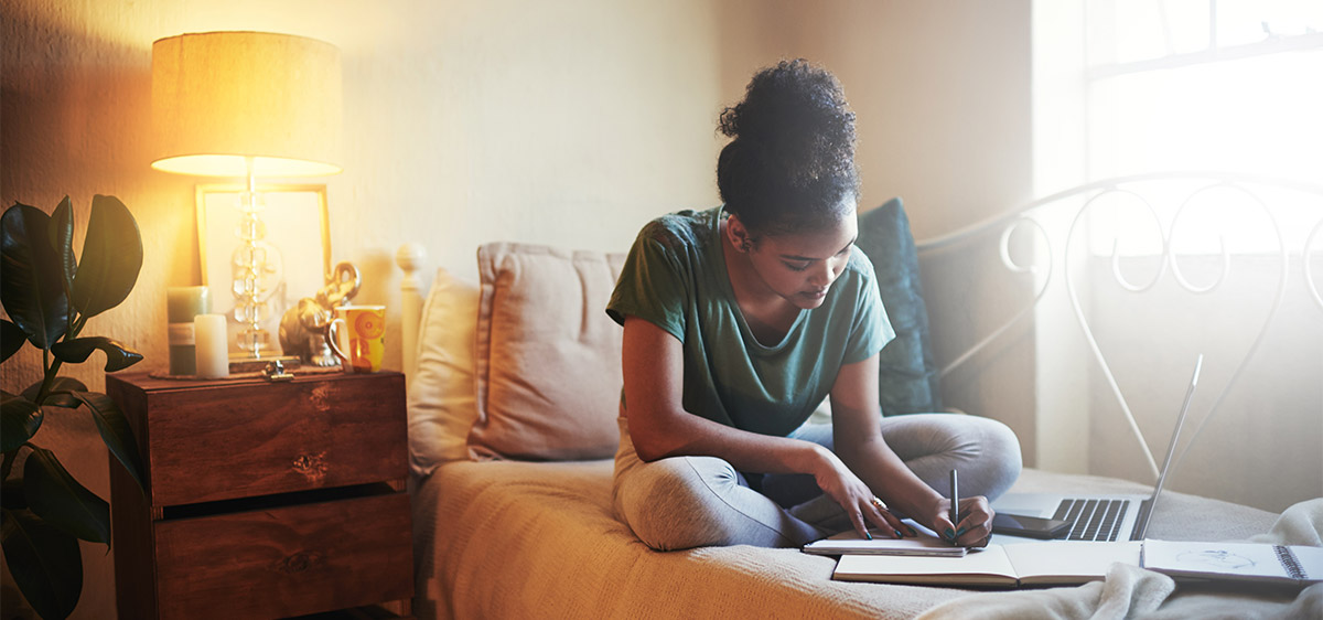 A student sits on a bed with laptop, notebooks.