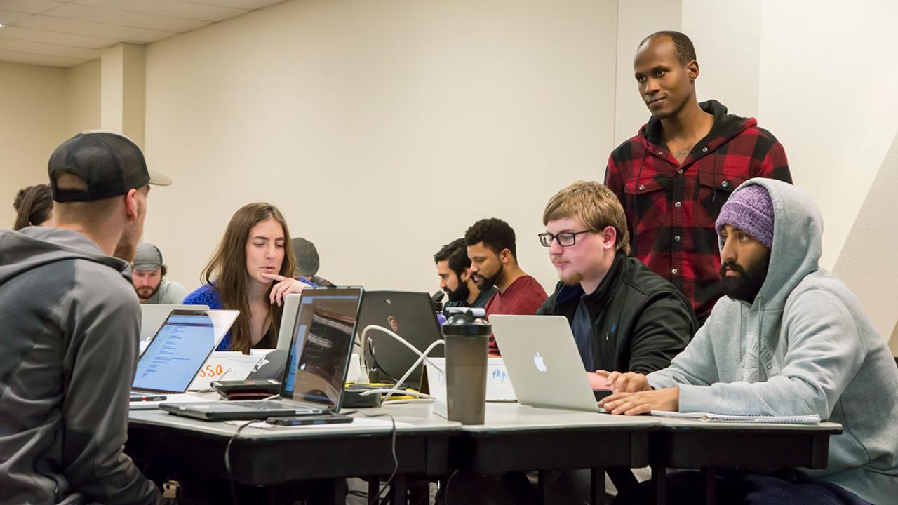 Instructor Nelly Sugira, standing to the right, helps students with a code-stringing exercise 