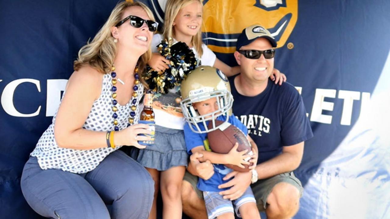 Photo: Jenny and Scott Seacrist and children in front of Aggie Athletics banner.