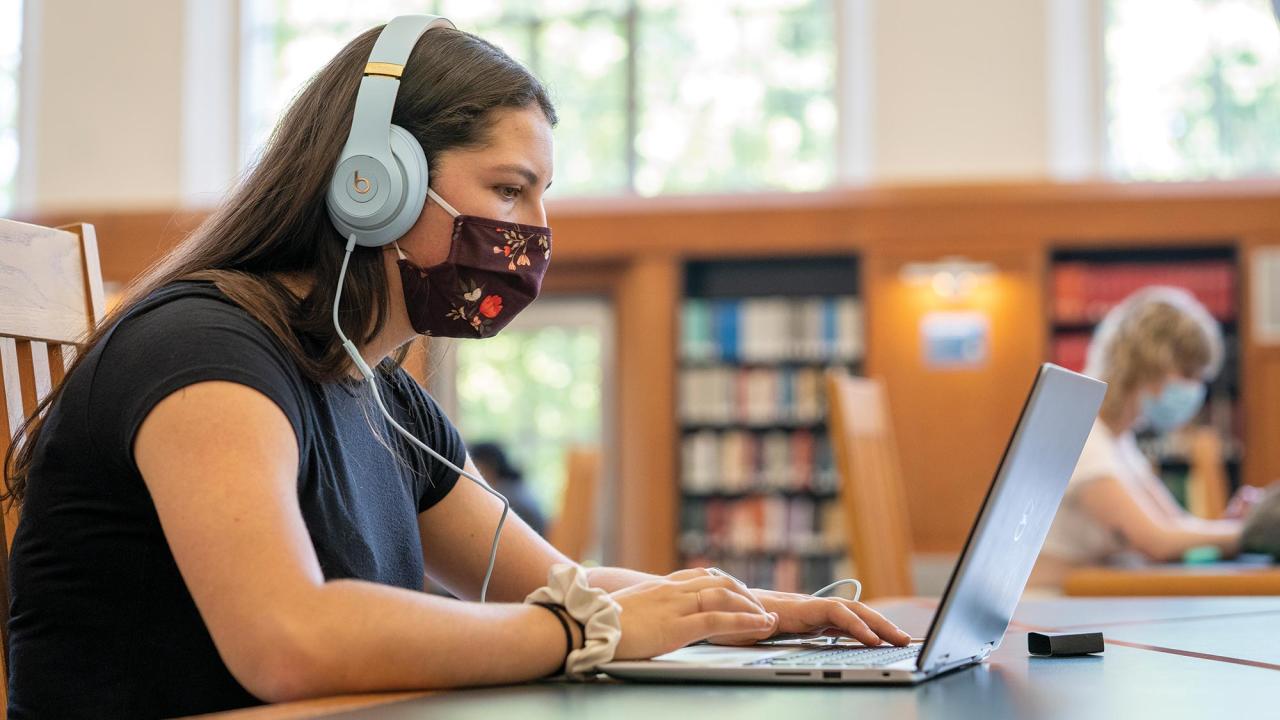 A woman student studies in the library. She is typing on an open laptop and wearing headphones and a face mask.