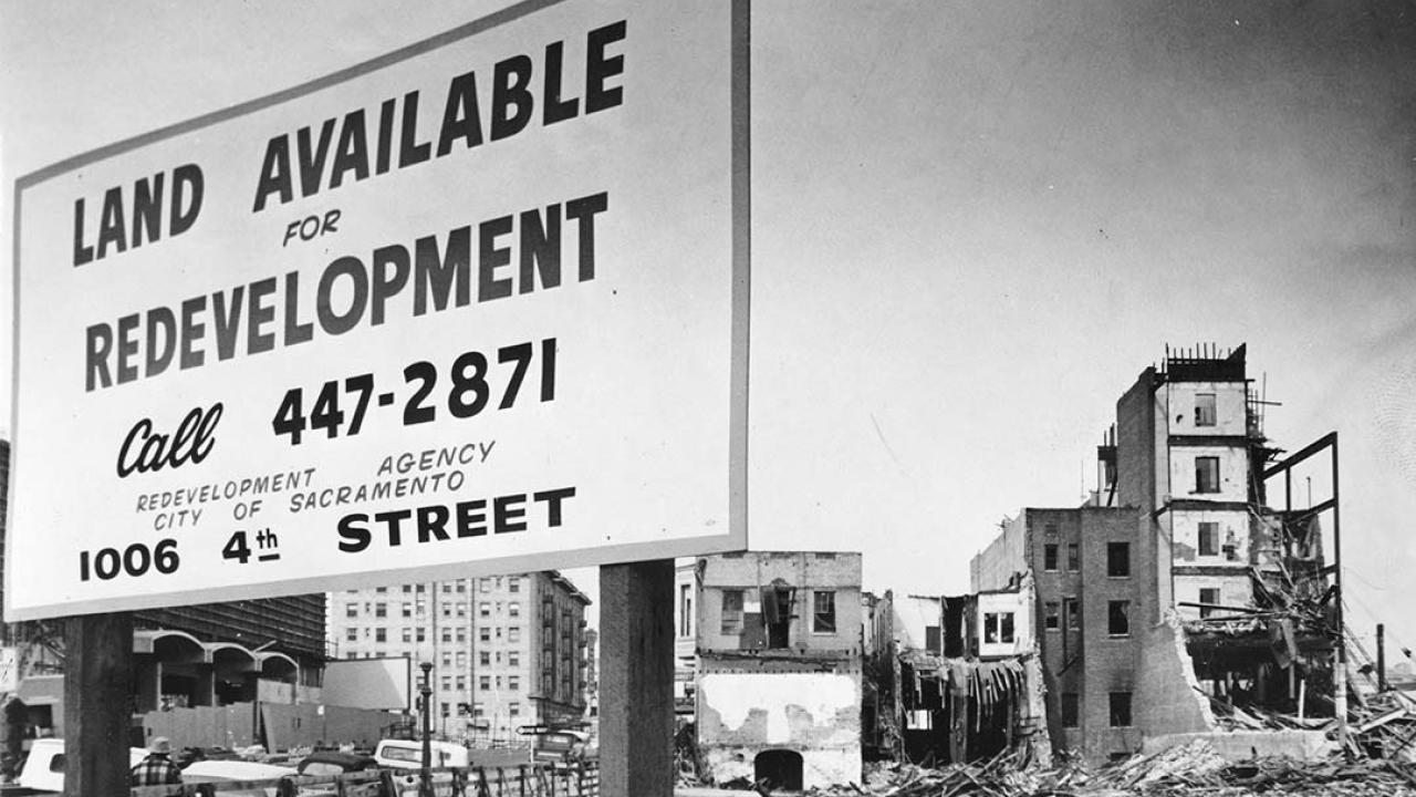 Black and white photo showing a large sign put up in front of a vacant lot. The sign reads "Land Available for Redevelopment" and gives a phone number and address. Buildings toward the back of the lot are undergoing various stages of demolition.