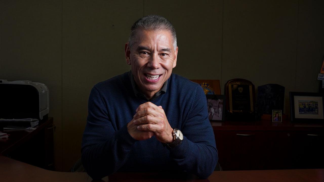 UC Davis Police Chief Joe Farrow sits behind his desk, smiling, with hands clasped together in front of him. He wears a blue shirt and silver watch.