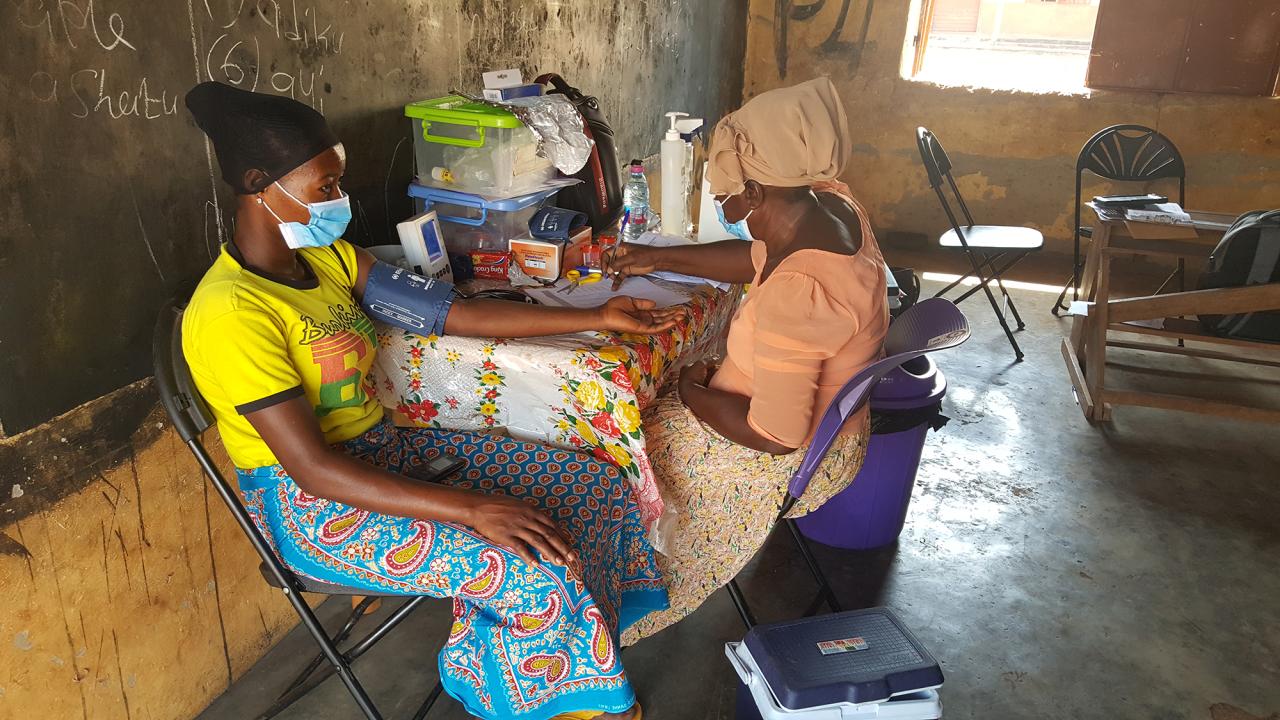Two African women sit at a table. The women at left wears a blue patterned skirt, yellow top and black headscarf, and a blood pressure cuff on her arm. The woman at right wears brown clothes and a headscarf and is making notes on a form. 