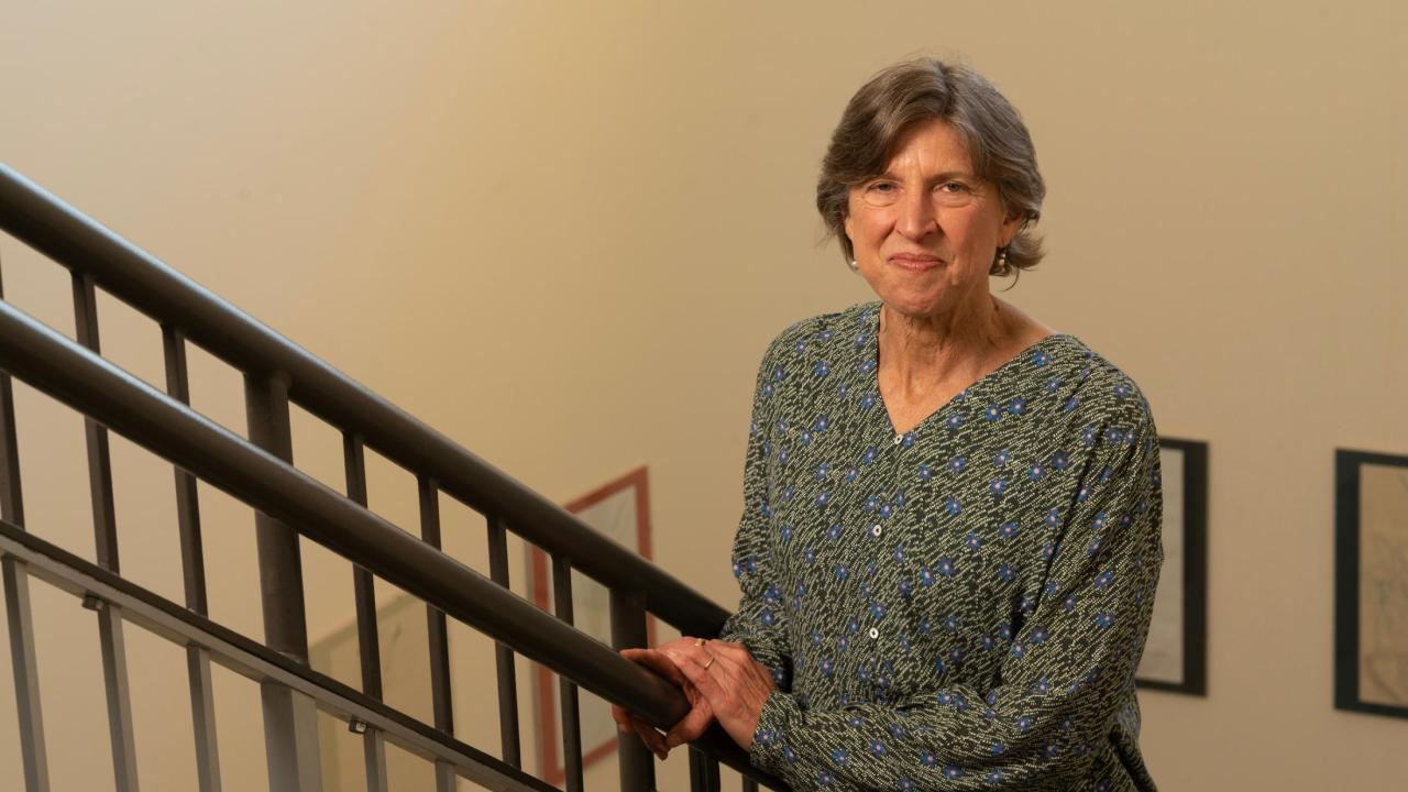 Woman in green patterned blouse on indoor staircase, holding railing and smiling towards the camera.