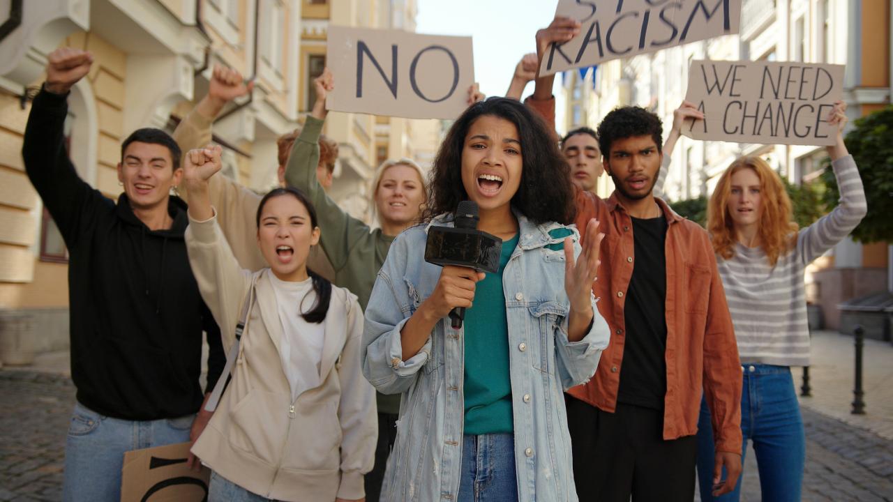 people holding protest signs in background as woman with microphone stands in front