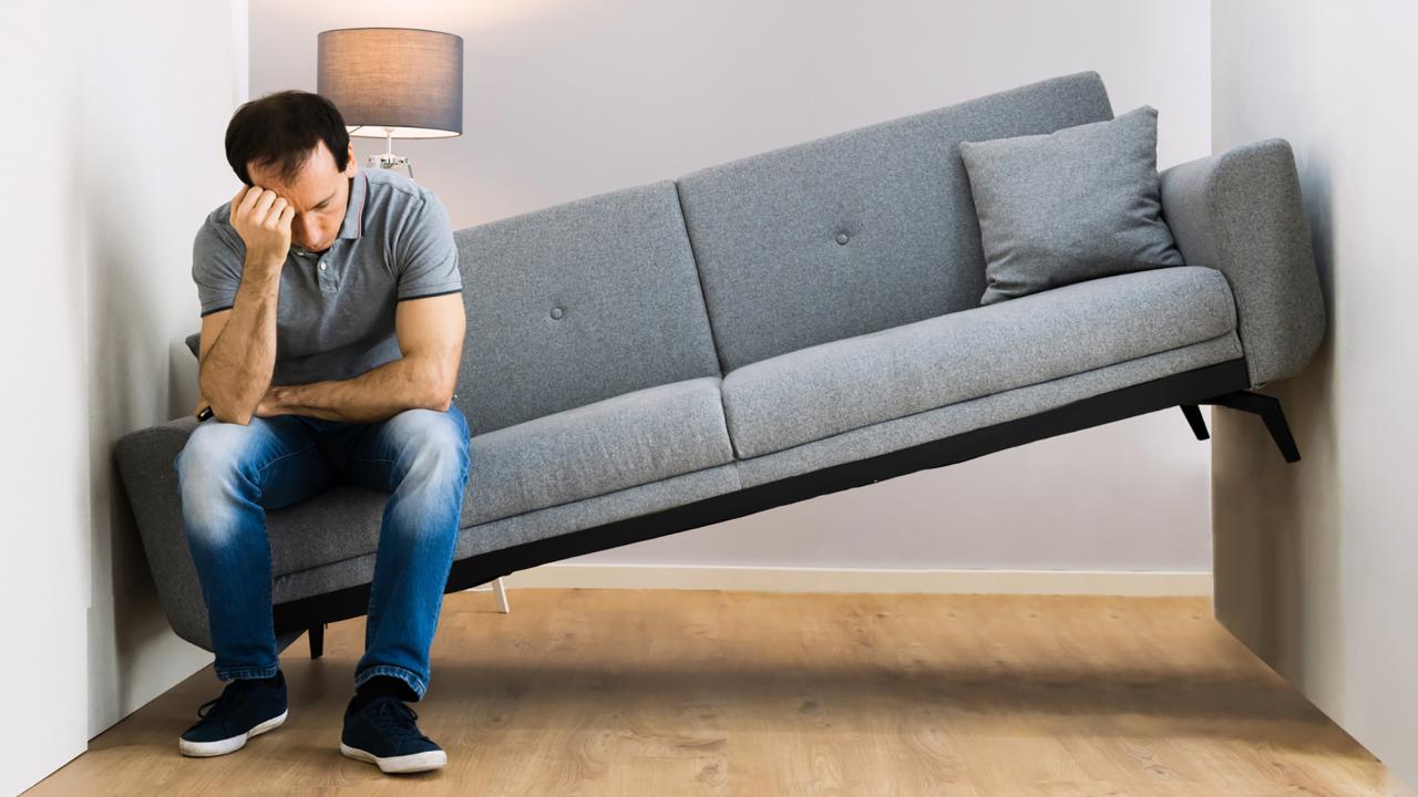 A despondent man sits at one end of a sofa wedged in a space too narrow for it. 