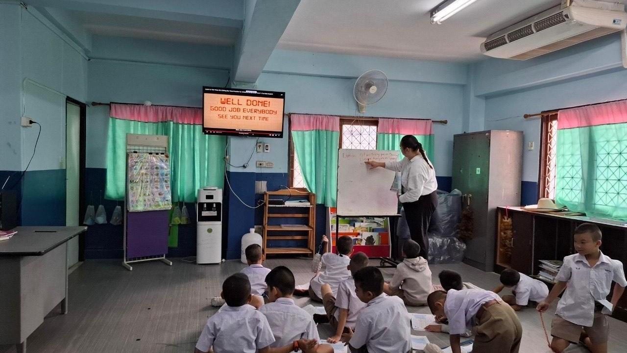 Ten young students in white shirts and tan pants, sit on a floor and face a teacher in a white shirt and black pants who writes on a whiteboard in a light blue classroom. 