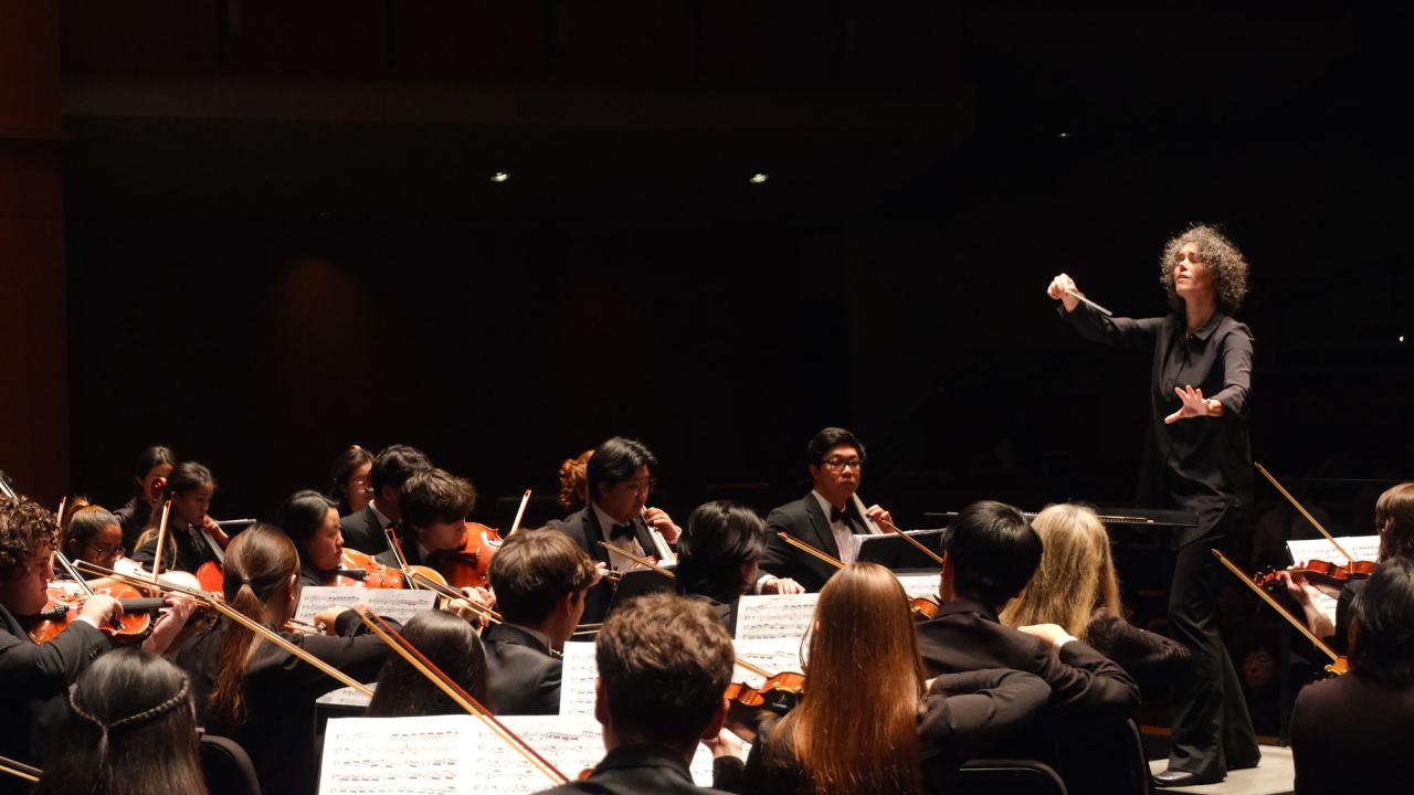 Orchestra on dark stage with conductor raising baton amid violinists under warm lights