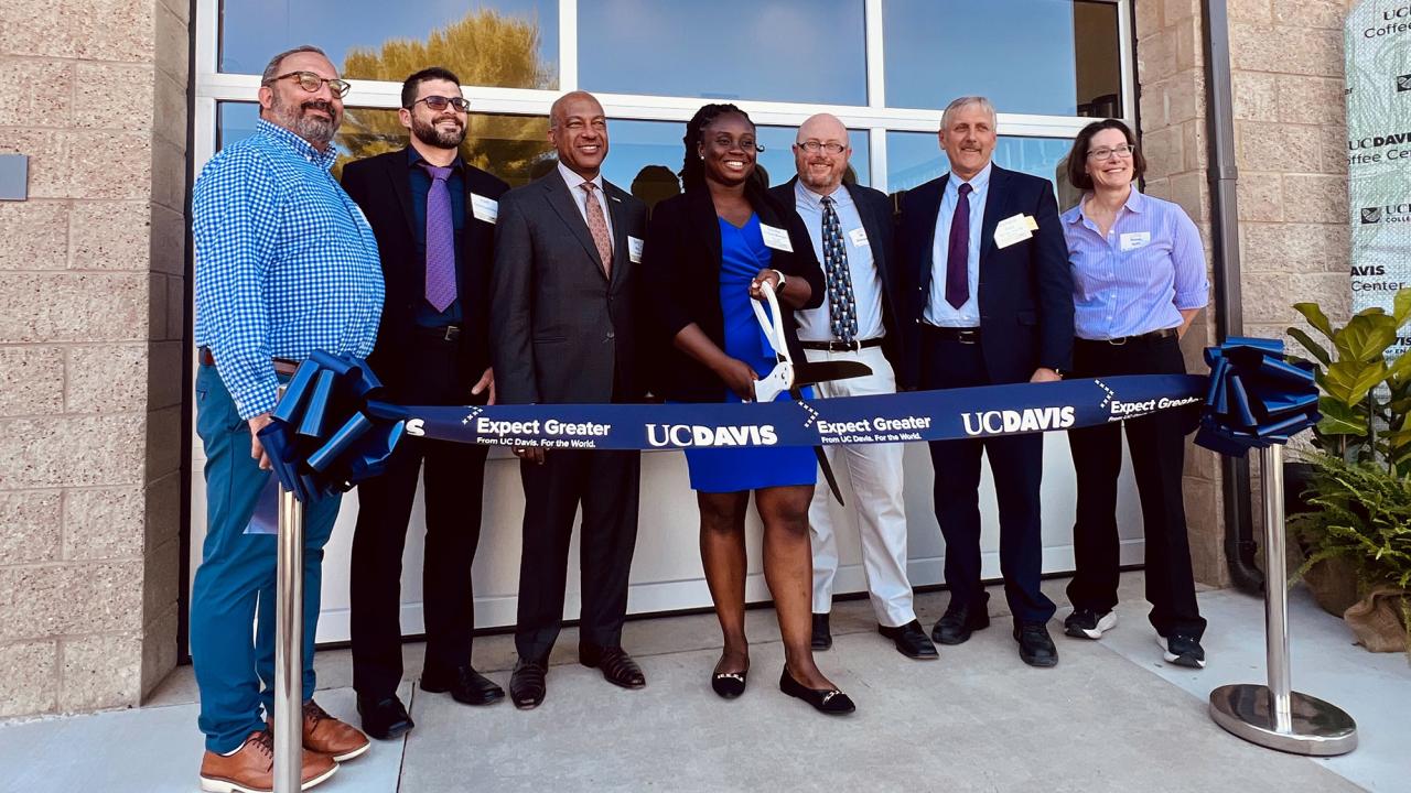 A group of men and women in semi-formal dress stand behind a blue ribbon held by stanchions. In the center, a black woman is about to cut the ribbon with large scissors. 