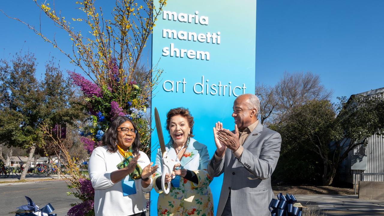Three people having cut a ribbon with a large scissors in front of Manetti Shrem sign