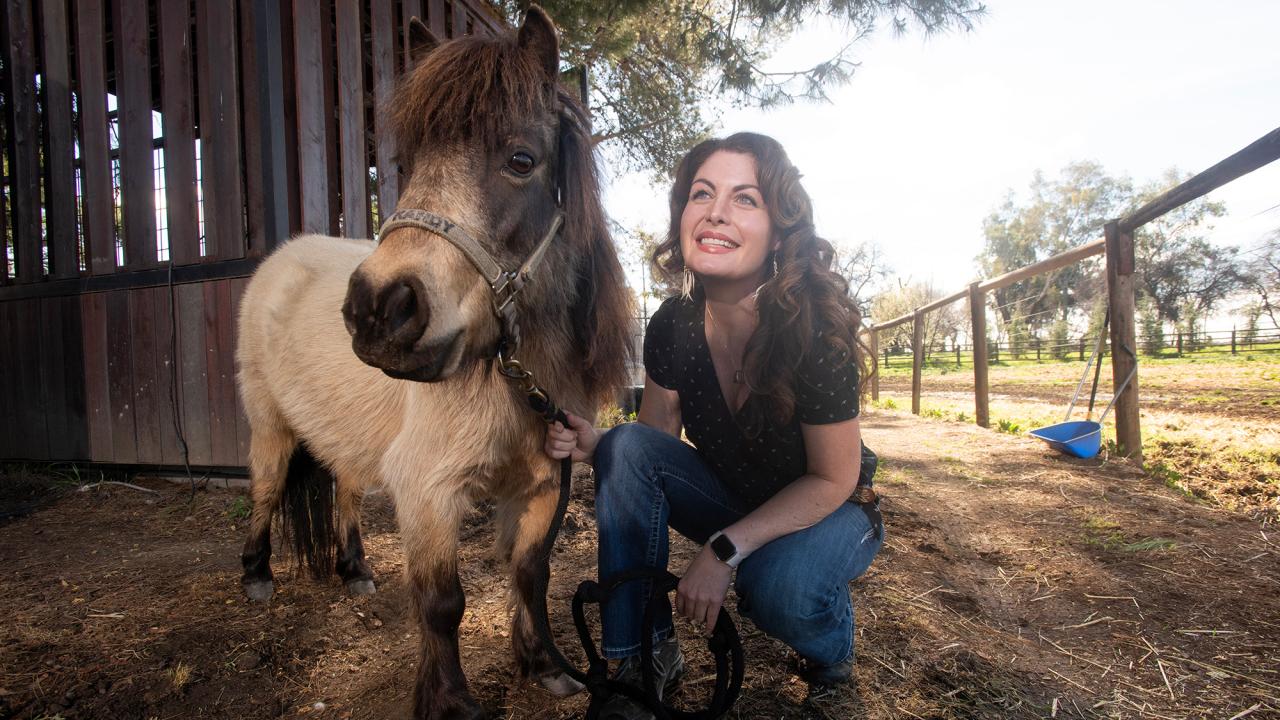 Woman crouching and smiling beside a small horse at a sunny farm