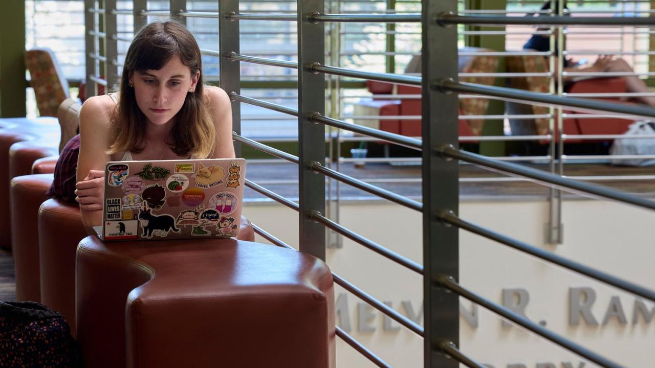 : A student lies on a padded bench inside the UC Davis Student Community Center and works on their laptop.