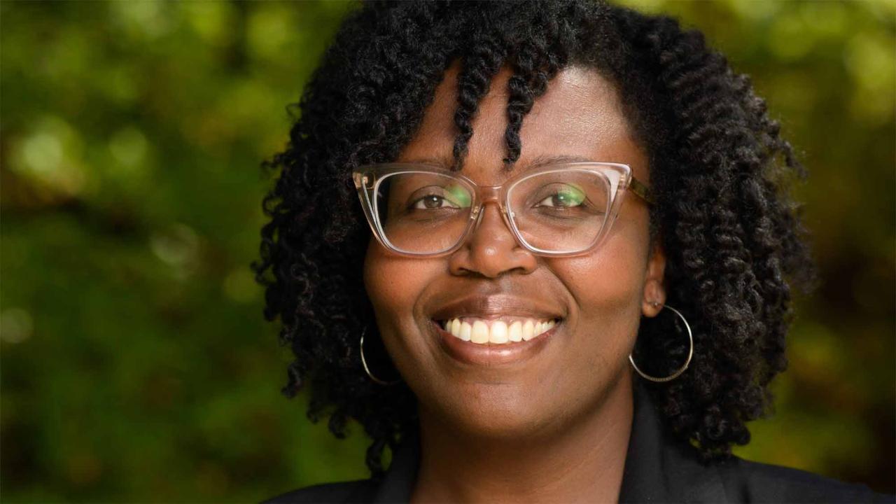 A woman wearing a black blazer smiles in front of a background of greenery