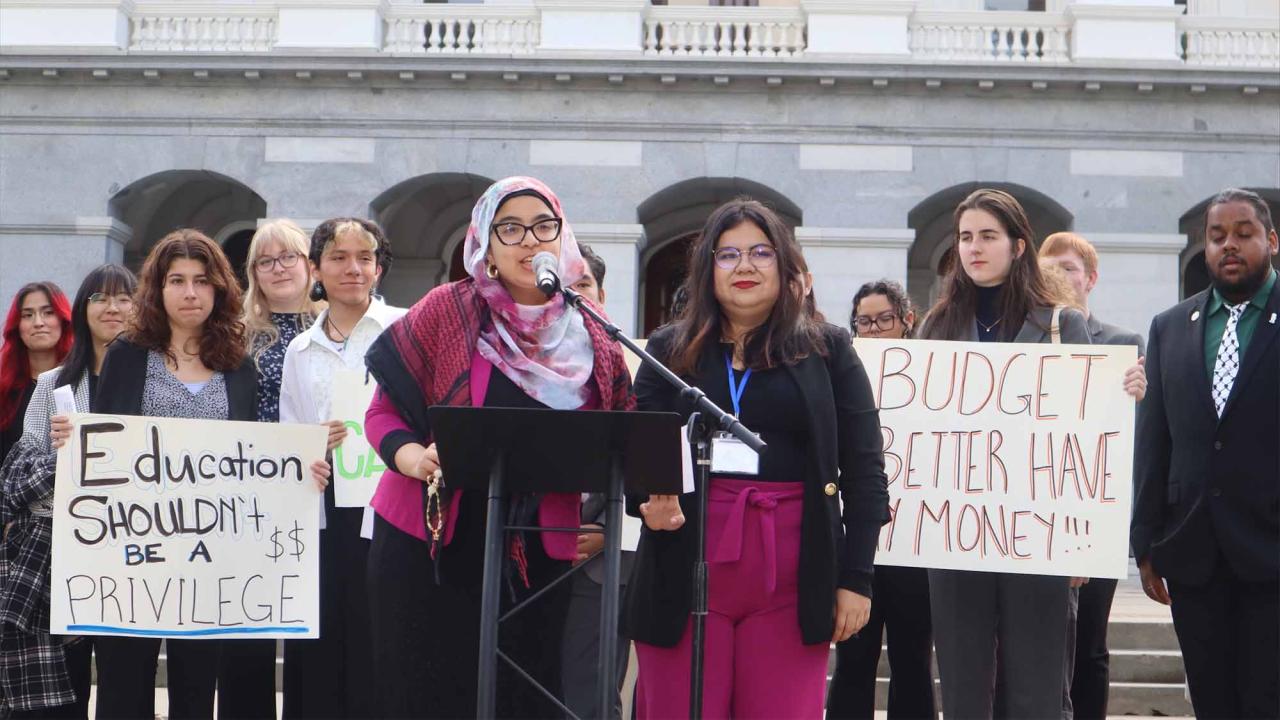 Aridin speaks at a podium in a hot pink outfit in front of a government building, surrounded by colleagues with signs that read “Education shouldn’t be a privilege” and “Budget better have my money.”