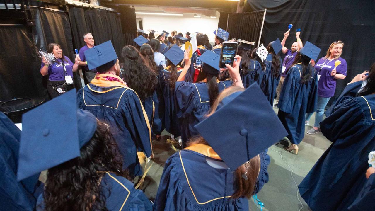 Graduates walk toward doorway in arena as volunteers in purple T-shirts cheer