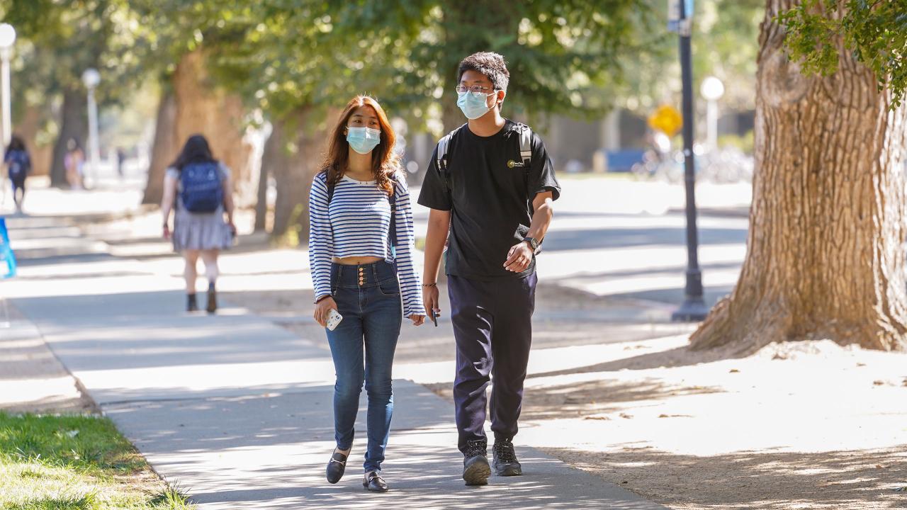 Students with masks walk through the sunlit UC Davis campus.