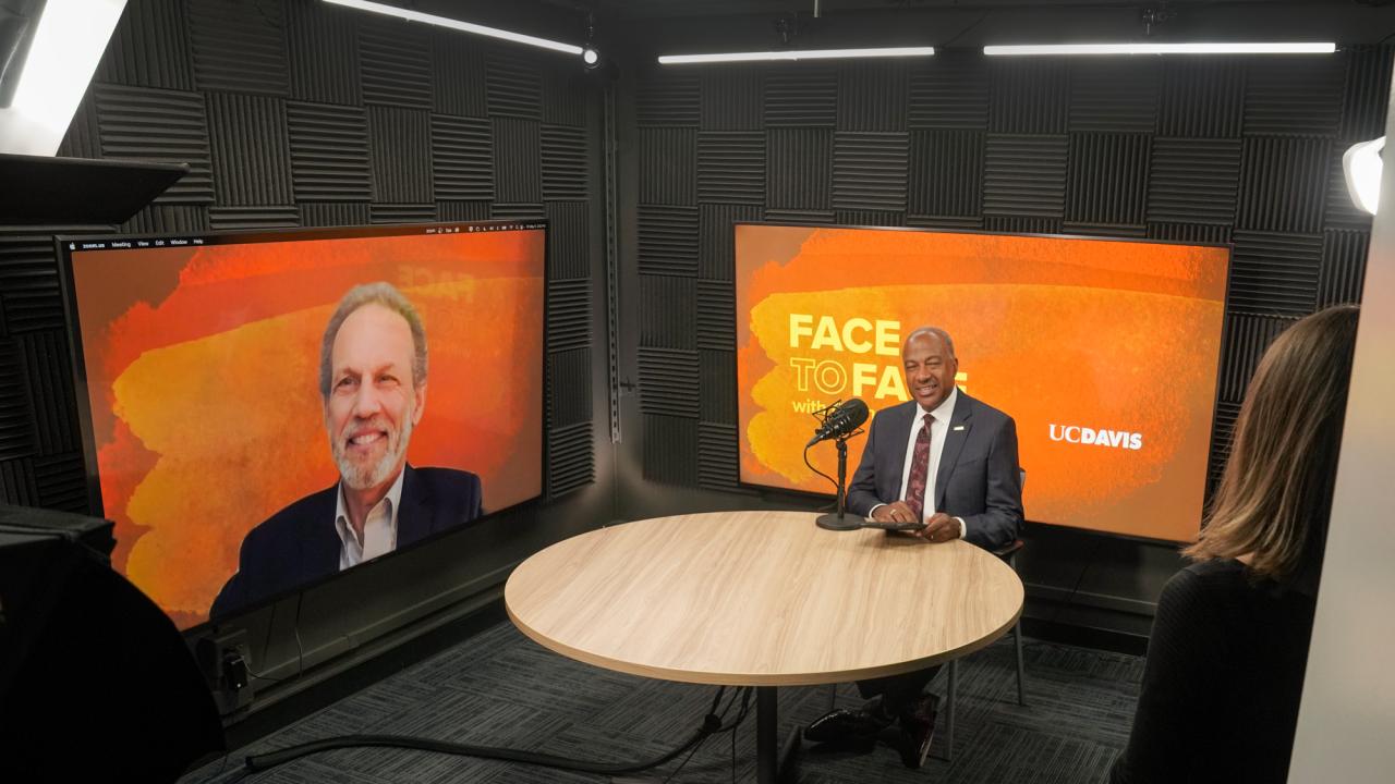 Chancellor Gary S. May, in suit and tie, at round table with microphone, in studio, for "Face to Face" recording with Bob Segar, on a monitor, appearing remotely from Boston