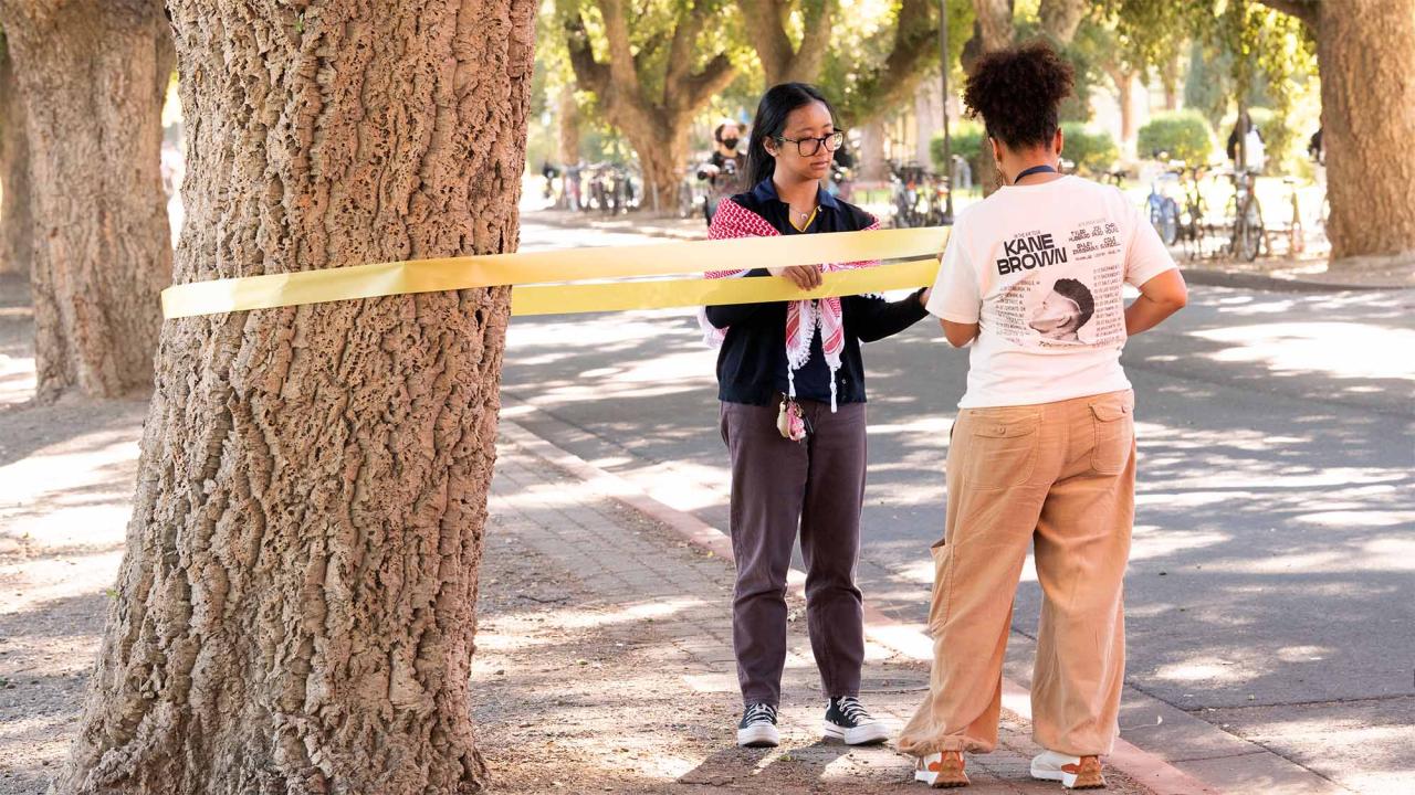 Two people tie yellow ribbon onto a tree