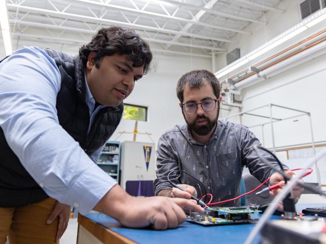 Two young men lean over a work table. Person on right, with short beard and glasses, is using a tool to test a circuit board as the person on left, with dark hair wearing a shirt and sleeveless vest, leans to examine it. 
