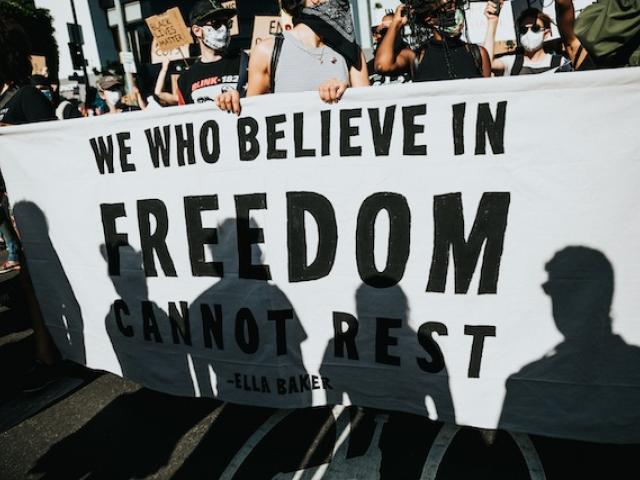 Marchers carry a large white banner with plain black lettering that says We who believe in freedom cannot rest.