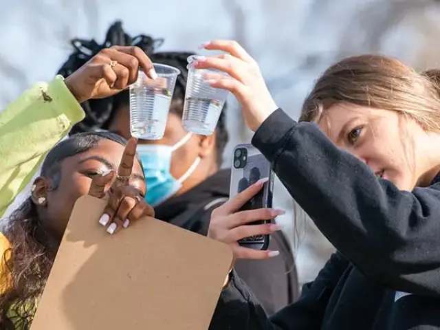 Three students closely examine clear plastic cups filled with water, holding them up to the light. One student points at a cup while holding a clipboard, another wears a face mask in the background, and a third uses a smartphone to take a photo or video. The scene takes place outdoors on a sunny day.