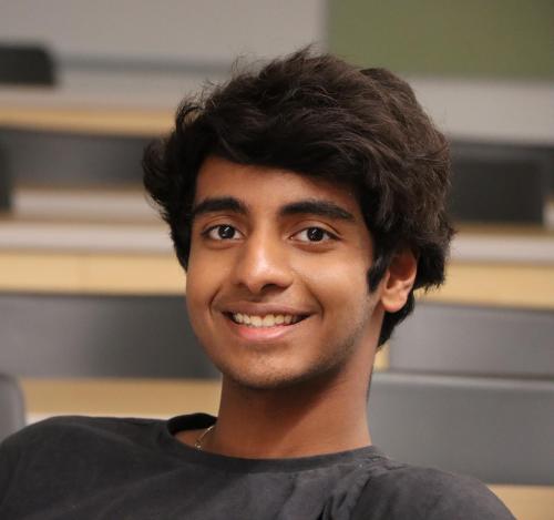 A student in a dark grey T-shirt, sits in a lecture hall, in front of a blurred background.