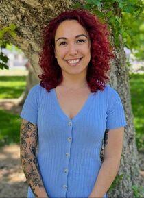 Head and shoulders of a smiling woman with red curly hair wearing a light blue short sleeved top with a tattoo on her right arm. 