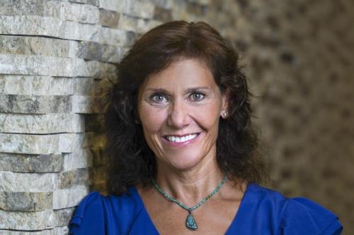 A woman with brown hair and wearing a blue shirt and a blue necklace smiles at the camera.