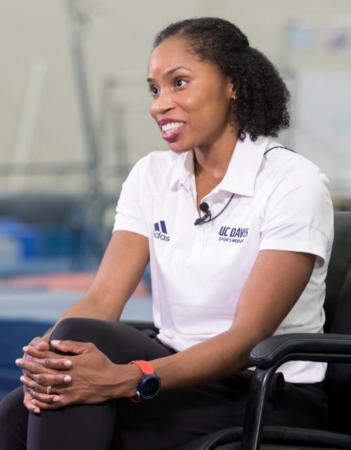 Female in white Aggie Athletics polo shirt, seated, hands clasped on knees