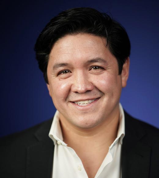 Smiling man with dark hair, wearing a black blazer over a white shirt, against a blue background.