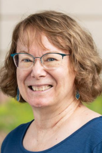 A smiling woman with curly brown hair and glasses, wearing a blue shirt, outdoors.