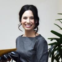 Rebecca. R. Hernandez smiles in headshot wearing gray top by plant