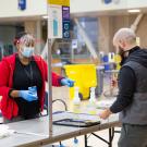 Woman behind plastic shield directs man at testing kiosk.