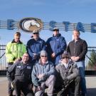 Seven men pose in front of Aggie Stadium gate and sign.