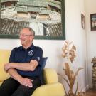 Ralph J. Hexter in Aggie polo shirt, seated on couch, under a painting.