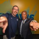 Aggie Football Coach Tim Plough and Chancellor Gary S. May point at the camera in energetic fashion while taking a photo in front of a wall with a UC Davis logo and gradient pattern. 