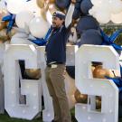 Three people celebrating by large white letters and gold, black, blue balloons outdoors