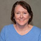 Smiling woman with short brown hair, wearing a blue sweater, in a library setting.
