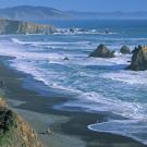 View of rocky coastline and beach to left with island rocks among the surf to right. 