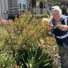 A large colorful bush outside a house fills the left and center of the screen. A woman with blonde hair wearing a blue jacket and hat is holding a branch of the bush and looking closely at it. 