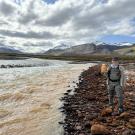 Scientist Taylor Evinger in green jacket and orange pack on rocky riverbank by muddy river and snow-capped mountains