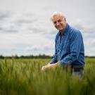 Smiling older man in blue shirt standing in green wheat field under cloudy sky. Jorge Dubkovsky, distinguished professor in the UC Davis Department of Plant Sciences, pictured here in the wheat fields at UC Davis.  (Jael Mackendorf / UC Davis) 