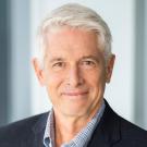 Professional headshot of gray-haired man in navy suit, slight smile, blurred office background
