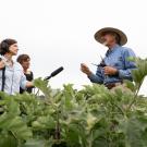 An interviewer holds a microphone up to another person standing in a farm field