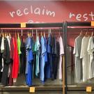 Clothes hanging on a rack, with the words "reclaim" and "restore" painted on the wall behind them.