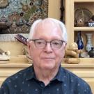 Michael Savageau at his home office, centered and starting at the camera with several personal artifacts resting on cupboards and shelves behind him.