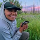 Sidney Woodruff in gray hoodie and NPS ballcap holds northwestern pond turtle while crouching beside a pond with pink sunset in background