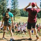 Two camp counselors stand on hill, shouting to group of campers assembled below.