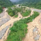 Aerial photo of a flooded river running through a narrow tropical valley. The river is muddy and fast-moving.