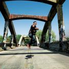 A man jumps in the air in the middle of a road that's on a bridge. The bridge is an old two-lane bridge with concrete trusses overhead. It's covered in graffiti.